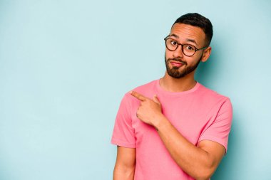 Mavi arka planda izole edilmiş genç İspanyol adam gülümsüyor ve boş alanda bir şeyler gösteriyor..