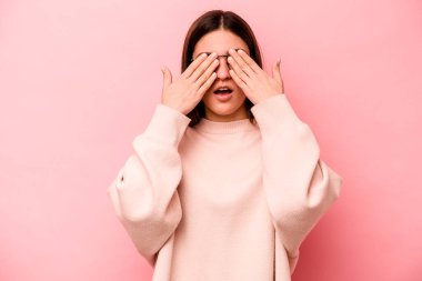 Pembe arka planda izole edilmiş genç beyaz kadın elleriyle gözlerini kapatmaktan korkuyor..