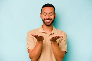 Genç İspanyol bir adam mavi arka planda izole edilmiş. Elinde avuç içi olan bir şeyle kameraya sunuyor..