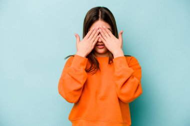 Mavi arka planda izole edilmiş genç beyaz kadın elleriyle gözlerini kapatmaktan korkuyor..