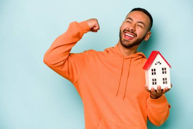 Young hispanic man holding a toy house isolated on blue background raising fist after a victory, winner concept.