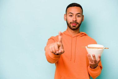 Mavi arka planda izole edilmiş erişte yiyen genç İspanyol kadın parmaklarıyla bir numarayı gösteriyor..