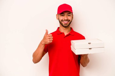 Beyaz arka planda izole edilmiş pizzaları tutan Latin kökenli genç teslimatçı gülümsüyor ve başparmağını kaldırıyor.
