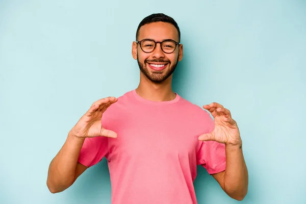 Jonge Spaanse Man Geïsoleerd Blauwe Achtergrond Met Iets Met Handpalmen — Stockfoto