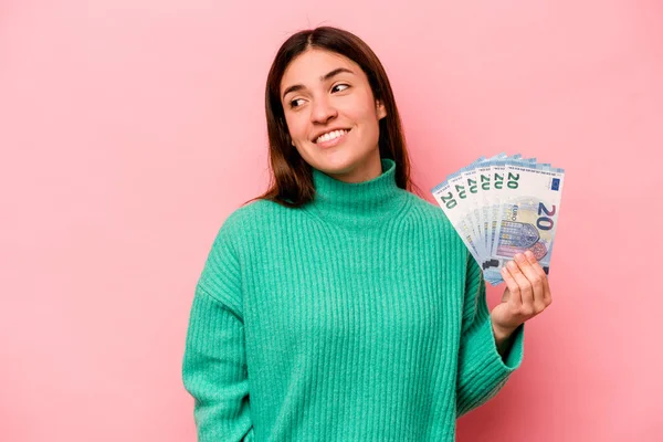 Young caucasian woman holding banknotes isolated on pink background looks aside smiling, cheerful and pleasant.