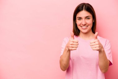 Pembe arka planda izole edilmiş genç beyaz kadın gülümsüyor ve başparmağını kaldırıyor.