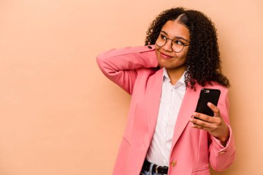 Elinde cep telefonu tutan genç Afrikalı bir kadın bej arka planda izole edilmiş kafasının arkasına dokunuyor, düşünüyor ve bir seçim yapıyor..