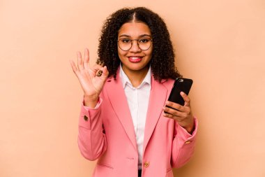 Elinde cep telefonu olan genç Afrikalı bir kadın bej arka planda izole edilmiş neşeli ve kendinden emin bir şekilde iyi niyet gösteriyor..
