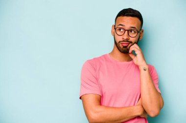 Genç İspanyol adam mavi arka planda izole edilmiş şüpheli ve şüpheci bir ifadeyle yana bakıyor..