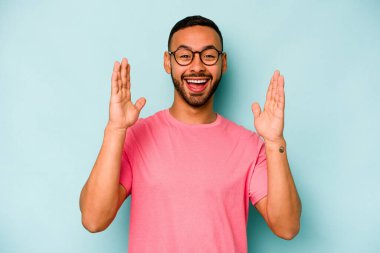 Genç İspanyol bir adam mavi arka planda izole edilmiş. Hoş bir sürpriz bekliyor. Heyecanlı ve el kaldırıyor..
