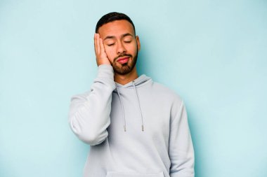 Genç İspanyol adam mavi arka planda izole edilmiş. Çok yorgun ve uykulu. Elini başının üstünde tutuyor..