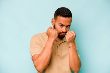 Latin kökenli genç bir adam mavi arka planda izole edilmiş. Yumruk atıyor, sinirleniyor, kavga ediyor..