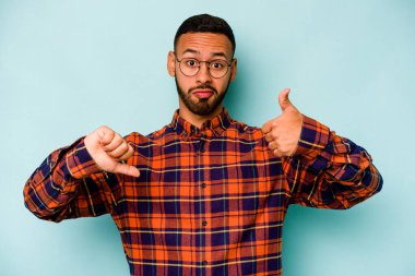 Latin kökenli genç adam mavi arka planda izole edilmiş, baş parmağını kaldırıyor, zor bir seçim konsepti.