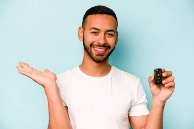 Young hispanic man holding car keys isolated on blue background showing a copy space on a palm and holding another hand on waist.