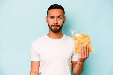 Mavi arka planda izole edilmiş bir paket cipsi tutan İspanyol genç adam kafası karışmış ve kendinden emin değil..
