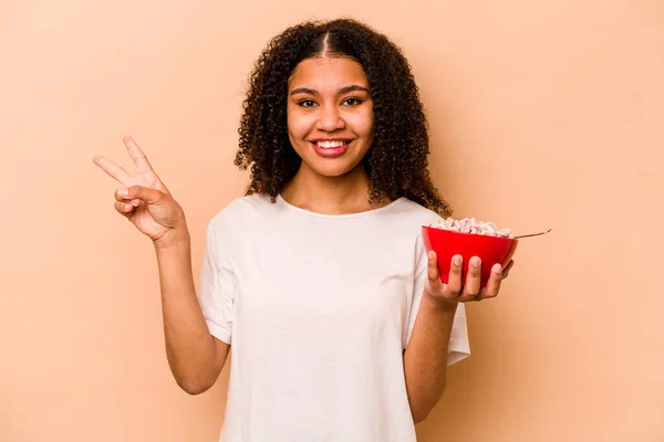 年轻的非洲裔美国女人拿着一碗谷类食品 在米色的背景下被隔离 快乐而无忧无虑 用手指展示着一个和平的象征 — 图库照片
