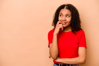 Genç Afrikalı Amerikalı kadın, bej arka planda izole edilmiş. Rahatlamış, fotokopi odasına bakan bir şey düşünüyor..