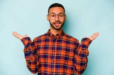 Genç İspanyol bir adam mavi arka planda izole edilmiş. Kafası karışmış ve omuz silkiyor. Kopya alanını tutmak için..