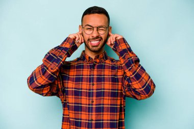 Mavi arka planda izole edilmiş genç İspanyol adam kulaklarını elleriyle kapatıyor..