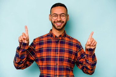 Mavi arka planda izole edilmiş genç İspanyol bir adam iki parmağı da boş bir alan gösteriyor..