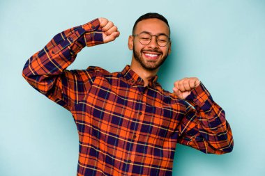 Genç İspanyol adam mavi arka planda izole edilmiş özel bir günü kutluyor, atlıyor ve enerjiyle kollarını kaldırıyor..