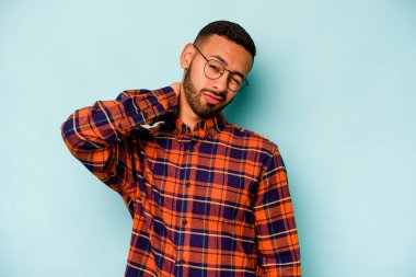 Genç İspanyol bir adam mavi arka planda izole edilmiş. Stres yüzünden boynu ağrıyor. Masaj yapıyor ve elle dokunuyor..