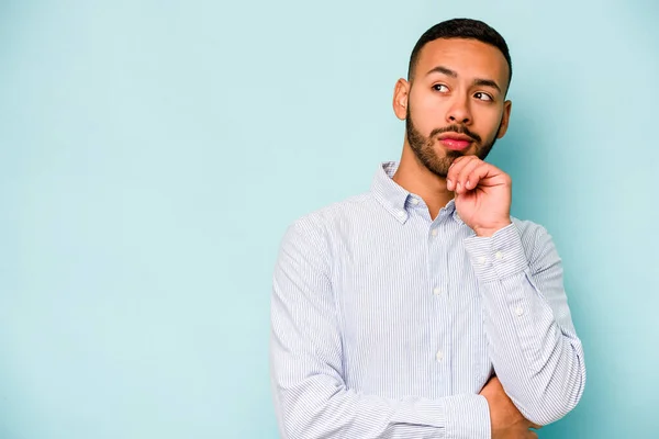 Young hispanic man isolated on blue background looking sideways with doubtful and skeptical expression.