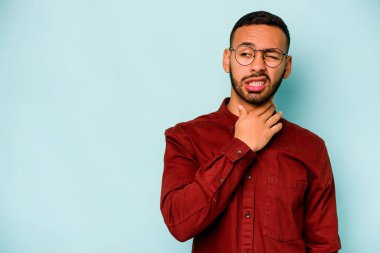 Genç İspanyol bir adam mavi arka planda izole edilmiş kafasının arkasına dokunuyor, düşünüyor ve bir seçim yapıyor..