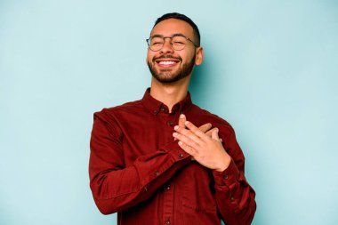 Mavi arka planda izole edilmiş genç İspanyol adam, avuçlarını göğsüne bastıran dostça bir ifadeye sahiptir. Aşk konsepti.