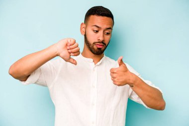 Latin kökenli genç adam mavi arka planda izole edilmiş, baş parmağını kaldırıyor, zor bir seçim konsepti.