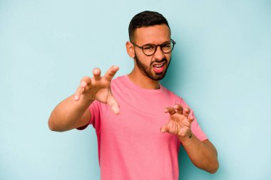 Mavi arka planda izole edilmiş genç İspanyol bir adam pençelerini bir kediyi taklit ederken gösteriyor, agresif bir hareket..