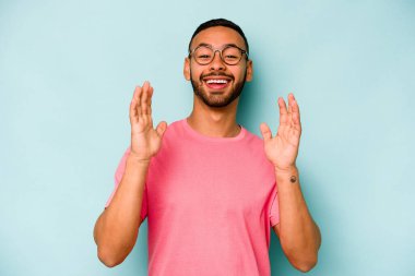 Mavi arka planda izole edilmiş genç İspanyol adam yüksek sesle gülüyor ve elini göğsüne koyuyor..