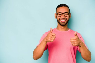 Genç İspanyol adam mavi arka planda izole edilmiş gülümsüyor ve başparmağını kaldırıyor.