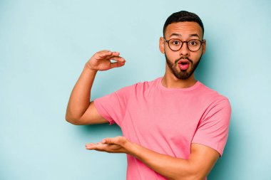 Mavi arka planda izole edilmiş genç İspanyol adam elleri arasında bir kopya alanı tutarken şok oldu ve hayrete düştü..