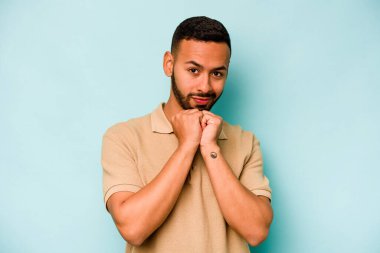 Mavi arka planda izole edilmiş genç İspanyol adam ellerini çenesinin altında tutuyor ve mutlu bir şekilde yan tarafa bakıyor..