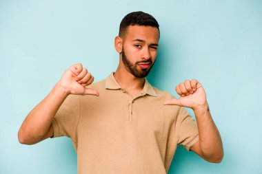 Mavi arka planda izole edilmiş genç İspanyol adam baş parmağını aşağı gösteriyor, hayal kırıklığı konsepti.