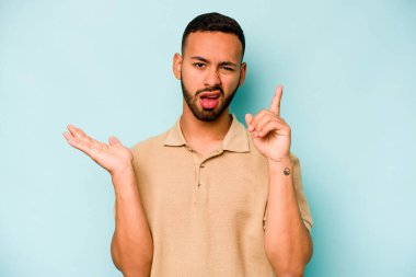 Genç İspanyol bir adam mavi arka planda izole edilmiş ve elinde bir ürün tutuyor..