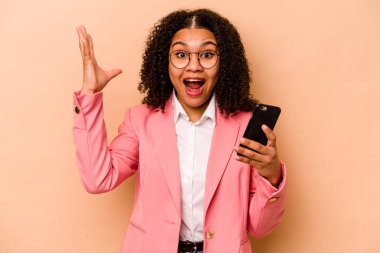 Genç Afrikalı Amerikalı bir kadın cep telefonunu bej arka planda tek başına tutuyor. Hoş bir sürprizle karşılaşıyor, heyecanlı ve el kaldırıyor..