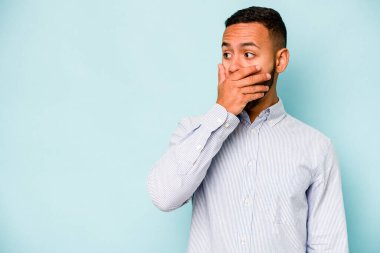 Genç İspanyol adam mavi arka planda izole edilmiş. Düşünceli bir şekilde bir fotokopi odasına bakıyor. Ağzını eliyle kapatıyor..