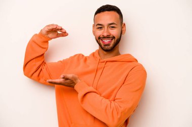 Beyaz arka planda izole edilmiş genç İspanyol adam işaret parmaklarıyla küçük bir şey tutuyor, gülümsüyor ve kendinden emin..