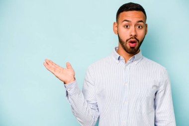 Mavi arka planda izole edilmiş genç İspanyol adam, avuç içinde kopya alanı tutmaktan etkilenmiş..