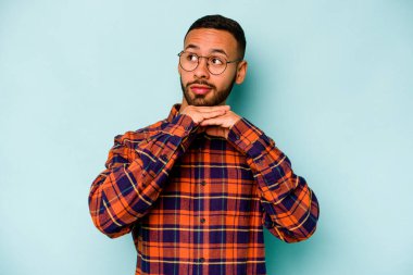 Mavi arka planda izole edilmiş genç İspanyol adam ellerini çenesinin altında tutuyor ve mutlu bir şekilde yan tarafa bakıyor..