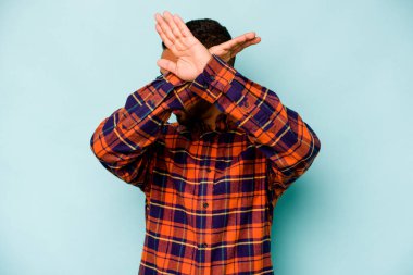 Genç İspanyol adam mavi arka planda izole edilmiş iki kolu çapraz tutuyor, inkar kavramı.