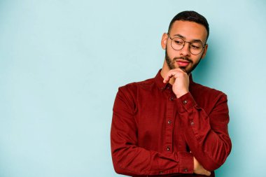 Genç İspanyol adam mavi arka planda izole edilmiş şüpheli ve şüpheci bir ifadeyle yana bakıyor..