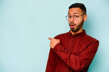 Mavi arka planda izole edilmiş genç İspanyol adam gülümsüyor ve boş alanda bir şeyler gösteriyor..