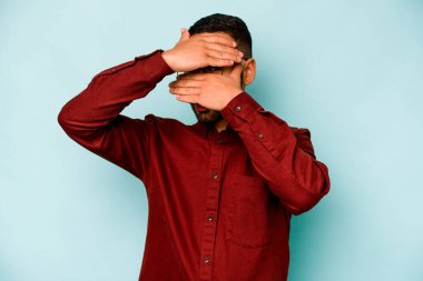 Mavi arka planda izole edilmiş genç İspanyol adam kameraya parmaklarıyla göz kırpıyor, yüzünü kapatmaktan utanıyor..