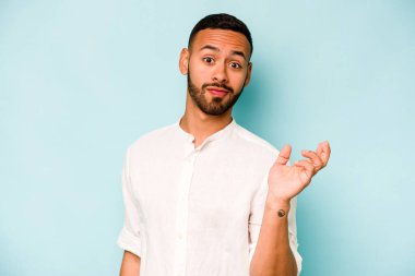 Latin kökenli genç bir adam, mavi arka planda izole edilmiş şüphe içinde ve sorgulama sırasında omuz silkiyor..