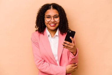 Elinde cep telefonu olan genç Afrikalı bir kadın bej arka planda izole edilmiş gülüyor ve eğleniyor..