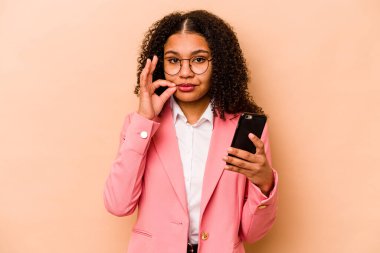 Cep telefonu tutan genç Afrikalı Amerikalı kadın bej arka planda izole edilmiş ve parmakları dudaklarında bir sır saklıyor..