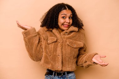 Genç Afrikalı Amerikalı kadın, bej arka planda izole edilmiş. Kafası karışmış ve omuz silkiyor..